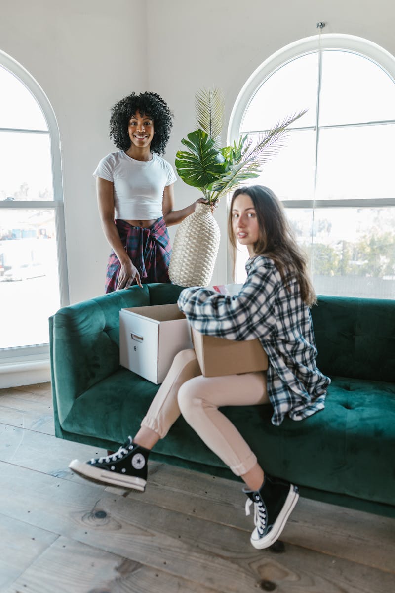 Two young women moving into a bright apartment, sitting on a green sofa with boxes. First Home.