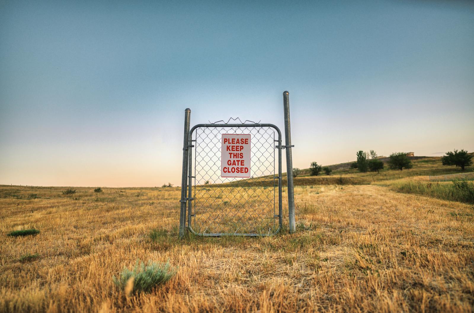 A steel gate with a warning sign stands in a vast deserted grass field under a clear sky.